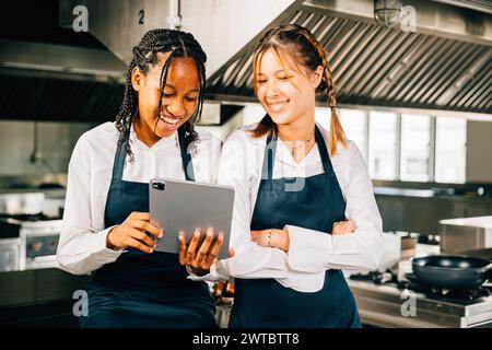 Two famous chefs in stainless steel kitchen discuss video blog using tablet. Professionals smiling Stock Photo