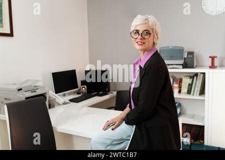 Middle-aged businesswoman, chic glasses, elegant blazer - poised and smiling in a sleek office environment - a portrait of experience and confidence. Stock Photo