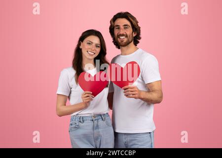 Happy couple in white t-shirts and jeans holding red heart-shaped paper cutouts Stock Photo