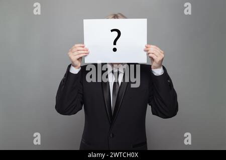 Formal anonymous person hiding his head behind white paper with question mark against gray studio wall background Stock Photo