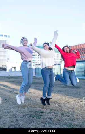 Three individuals woman, dressed in jeans and sweaters, are captured mid-jump in an outdoor field. Arms extended, they express freedom and joy. Urban Stock Photo