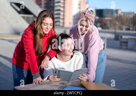 Three young women leaning over a notebook on a table outside, in focused discussion, with one looking up and smiling, in a sunny urban setting Stock Photo