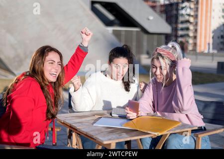 Three young women gather around a tablet and documents, engaged in a group study session outdoors. They're focused yet relaxed, with an urban backdrop Stock Photo