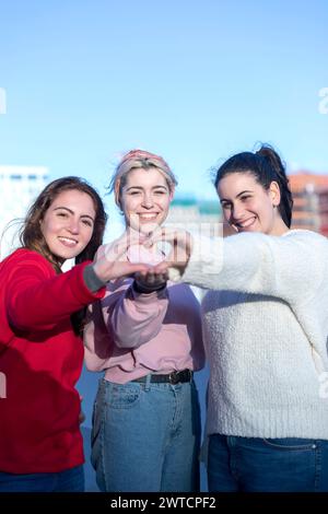 Three smiling young women outdoors join hands to form a heart shape, symbolizing friendship and love, with one in red, another in pink, and the third Stock Photo