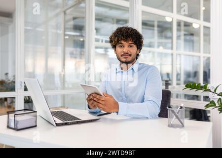 A focused young man in a blue shirt engaged in multitasking with a laptop and tablet at a bright office desk. Stock Photo