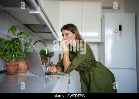 Thoughtful woman stands in front laptop table kitchen, focused on reading information from screen. Stock Photo