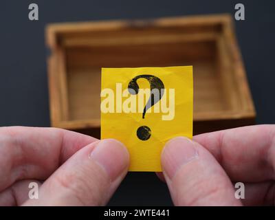A man holding a piece of yellow paper with the question mark on it in his hands over a small empty wooden box. Close up. Stock Photo
