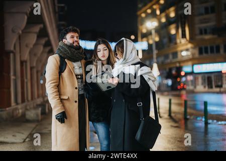 A trio of young adults engage in a discussion on an urban street at night, possibly collaborating on a creative project or work-related task. Stock Photo