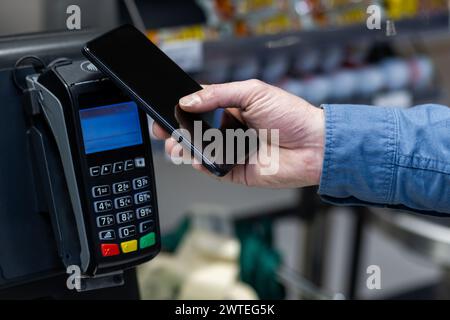 Close-up of a male hand holding a smartphone to a card reader for mobile payment in a retail store setting. Stock Photo