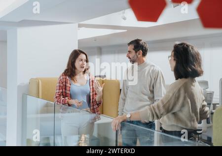 A group of three young people are standing and talking in a modern office setting, exuding a casual yet engaged atmosphere during their discussion. Stock Photo
