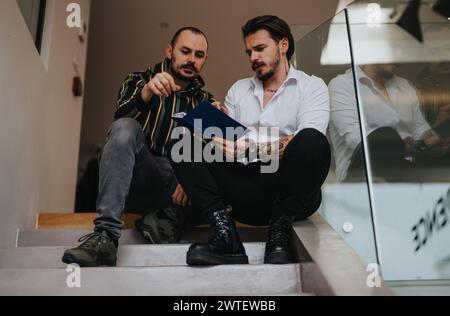 A professionally dressed man with tattoos collaborates with a colleague, going over a blue folder's contents on an indoor staircase. Stock Photo