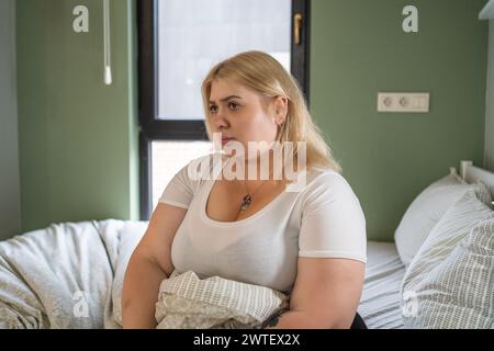 Lonely Plus-size woman sits on bed, looking weary, embodying feelings of life burdens and problems. Stock Photo