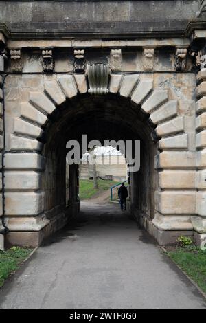 Person walks under a historic stone arch in a serene park setting Stock Photo