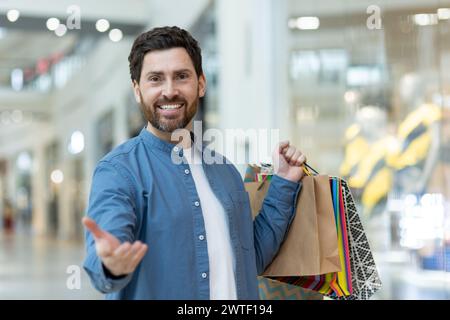 Smiling bearded man holding shopping bags extends a hand in greeting at a bright mall setting. Stock Photo