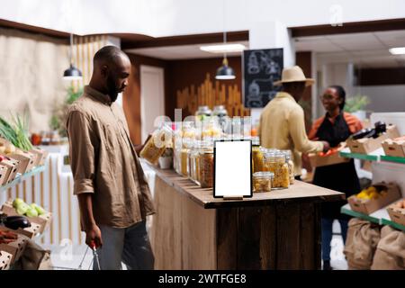 In an eco friendly store, a detailed look of a tablet with an isolated chromakey mockup template is shown. Close-up of digital device with blank white screen display, positioned near food glass jars. Stock Photo