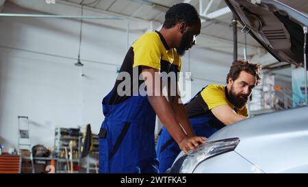 Mechanics in auto repair shop working together on fixing car, using tablet and laptop. Coworkers collaborating on servicing broken vehicle, checking for defective components using electronic devices Stock Photo