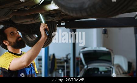 Precise mechanic underneath vehicle on overhead lift in garage, checking components. Employee using work light to make sure automotive underbody is in perfect condition, doing close examination Stock Photo