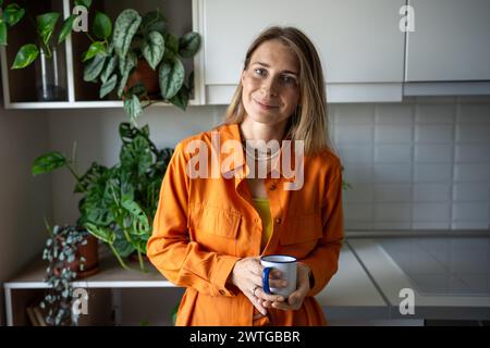 Smiling relaxed woman plant lover in orange dress holding mug in cozy kitchen, look at camera Stock Photo