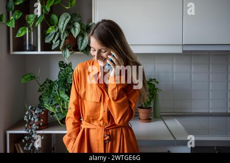 Smiling pleased woman enjoying pleasant conversation talking on mobile phone on kitchen at home.  Stock Photo