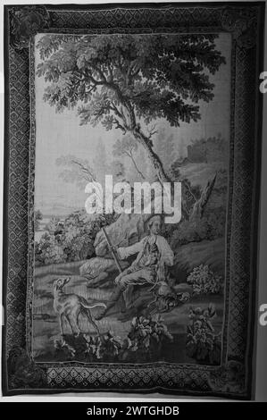 Hunter resting with dogs. unknown c. 1750-1800 Tapestry Dimensions: H 8'10' x W 5'9' Tapestry Materials/Techniques: unknown Culture: French Weaving Center: Aubusson Ownership History: French & Co. purchased from Mrs. H. S. Vanderbilt, invoiced 4/21/1942; sold to Dr. Nicolas Karger 1/29/1959. In landscape with trees & flowering plants, a hunter sits on grassy mound holding a fowling-piece & patting a dog, next to dead game & with additional dog in foreground; buildings on top of hill in distance (R) (BRD) picture-frame border with trellis pattern enclosing rosettes, & scrolled acanthus leaves i Stock Photo