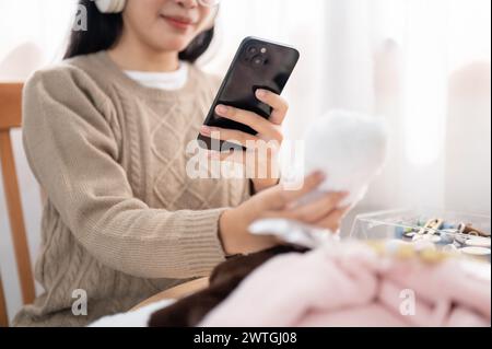 A cropped image of a positive Asian woman using her smartphone, taking a picture of her handmade plushie at her sewing table at home. Stock Photo
