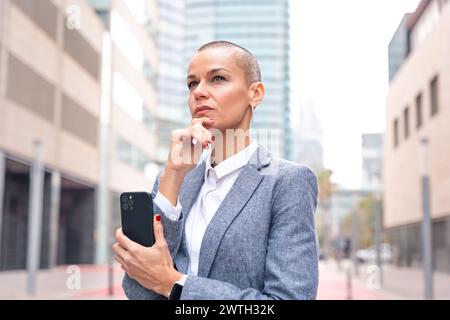 Caucasian business woman with pensive face using an app on her smartphone outdoors. Stock Photo