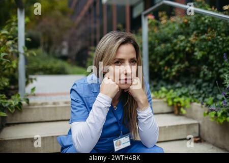 Female doctor feeling overwhelmed at work, sitting on stairs. Healthcare workers with stressful job, feeling exhausted. Burnout syndrome for doctors Stock Photo