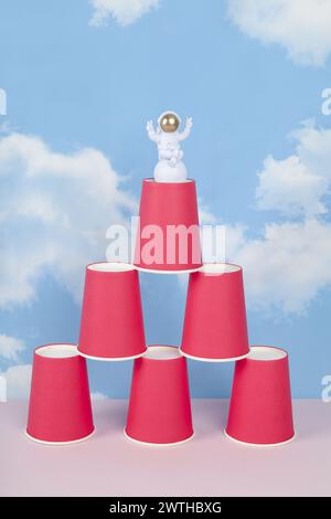 a white astronaut figurine with gold helmet on a pyramid of red paper cups, set against a background of blue summer sky with white clouds.Vivid color Stock Photo