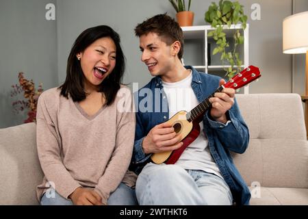 Happy young diverse couple singing a song playing guitar on sofa in living room at home. Lovely young adult woman and man having fun with ukulele in Stock Photo