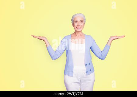 Beautiful nice cheerful smiling gray-haired old lady wearing casual, showing two options with palms. Isolated over pastel violet purple background Stock Photo