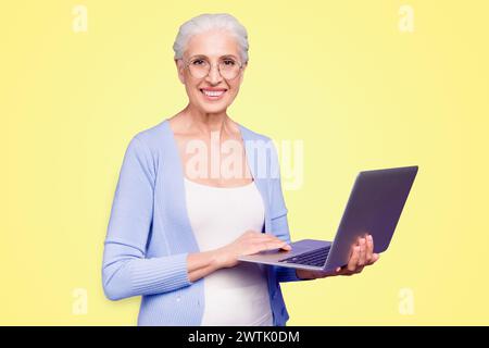 Grey haired old nice beautiful cheerful smiling woman wearing spectacles holding laptop learning online course. Isolated over violet purple background Stock Photo