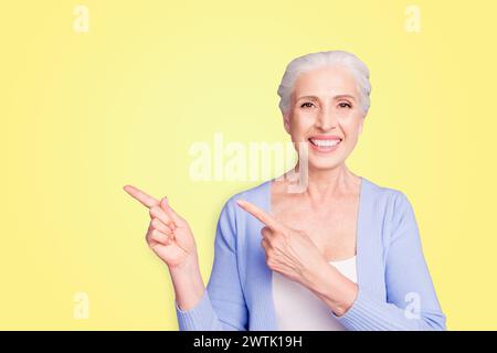 Beautiful nice cheerful smiling gray-haired old lady wearing casual, showing pointing way with two fingers. Copy space. Isolated over pastel violet Stock Photo