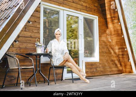 Barefoot woman in bathrobe sits on terrace of country wooden cabin. Concept of renting of tiny house to retreat Stock Photo