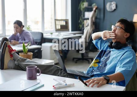 Side view portrait of Asian young man yawning while working with laptop in office feet up on table copy space Stock Photo