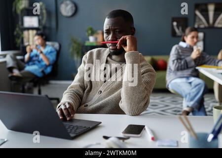Portrait of bored African American man playing with pencil while working at desk in office and looking at laptop screen Stock Photo