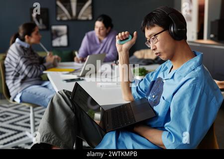 Side view portrait of young Asian man looking at laptop screen in office while casually sitting at workplace with hand expander copy space Stock Photo