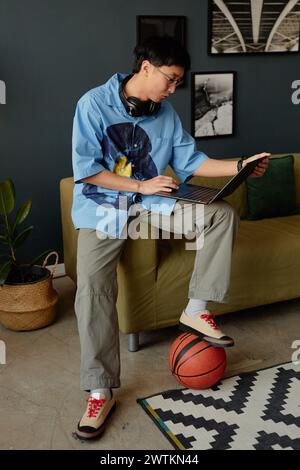 Vertical full length portrait of Asian young man using laptop at home or in office lounge and playing with basketball Stock Photo