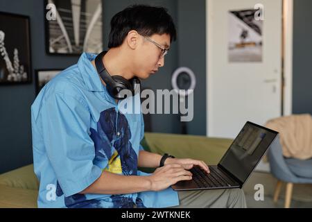 Side view portrait of Asian young man using laptop and wearing big headphones while working on IT and web design copy space Stock Photo