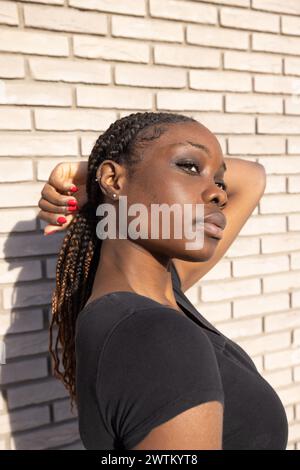 Captured in a moment of quiet repose, this photograph features a young African woman with her eyes gently closed, radiating tranquility. The sunlight caresses her skin, subtly highlighting her facial structure against the stark white bricks behind her. Her hands, adorned with red nail polish, are raised in an elegant gesture to her hair, braided in a style that speaks of cultural heritage. This casual yet striking pose, combined with the minimalist black t-shirt, creates a juxtaposition of simplicity and complexity. The softness of the light and the muted background amplify the introspective a Stock Photo