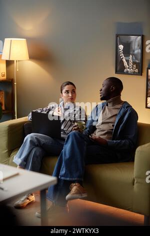 Vertical full length portrait of man and woman discussing project ideas while sitting on couch in office and holding laptop in low light Stock Photo