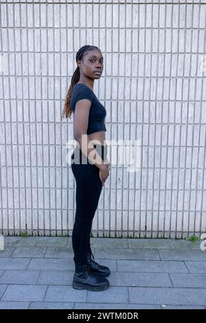 This image captures a young African woman standing confidently against a light-colored brick wall. Her posture is relaxed yet assertive, as she gazes directly into the camera with a neutral expression. She wears a casual black crop top and dark jeans, complemented by black lace-up boots. Her long hair is styled in neat, slender braids that hang down her back. The simplicity of the backdrop highlights her presence and attire, suggesting themes of urban fashion and youth culture. Confident Young Woman Against Brick Wall. High quality photo Stock Photo