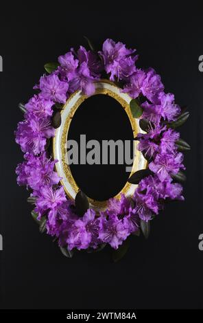 An oval picture frame surrounded by purple rhododendron flowers on a blank black background. Stock Photo