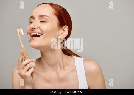 excited woman with red hair holding bamboo toothbrush with toothpaste on grey backdrop, dental care Stock Photo