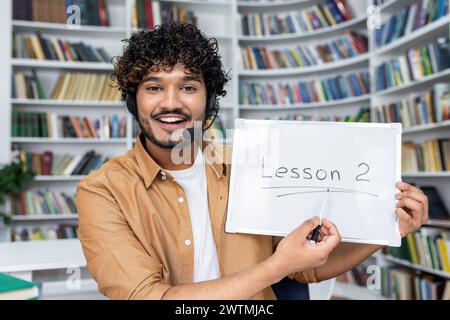 Young arab tutor in headset with microphone holding drawing board with lesson 2 inscription while smiling at camera. Positive man pointing with pen to tablet on background of bookshelves. Stock Photo