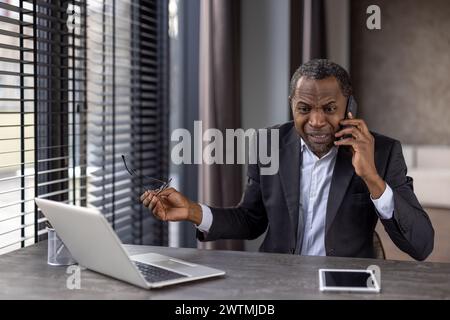 Furious diverse male in formalwear holding eyeglasses while having unpleasant phone conversation. Angry distant worker hearing bad breaking news while sitting by desktop with portable computer. Stock Photo