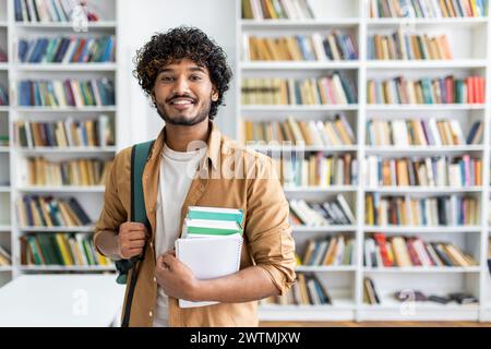 Happy unshaved man wearing backpack and holding stack of books while smiling at camera at bookstore. Optimistic classman making purchases for beginning of new academic semester in college. Stock Photo