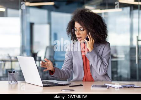 Terrified female talking by mobile phone while looking at portable computer screen with rounded eyes. Furious black woman getting desperate while receiving bad urgent news by business partner. Stock Photo