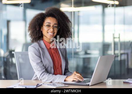 Attractive african american freelancer smiling at camera while keeping hands on portable computer keyboard. Successful interior designer enjoying productive busy day in modern shared coworking. Stock Photo