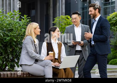Company of coworkers in formalwear resting outside of corporate building in friendly atmosphere. Two men standing by couple of women sitting on bench with coffee cup and laptop during lunch break. Stock Photo