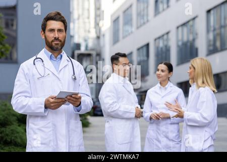 Experienced caucasian surgeon with stethoscope over neck standing with back to three interns talking about practice. Medical university student gathering in front of clinic in professional uniform. Stock Photo